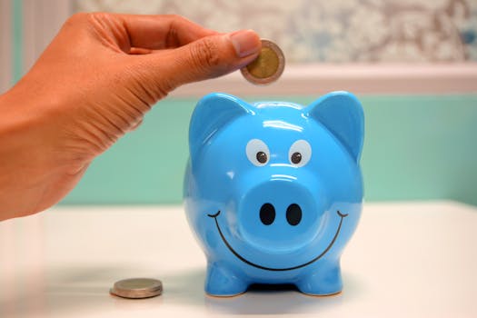 pexels-photo-1602726-1602726-1 Hand inserting a coin into a blue piggy bank for savings and money management.