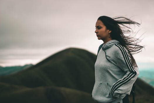 Profile view of a woman in an adidas hoodie with windswept hair standing by a mountain during daylight.