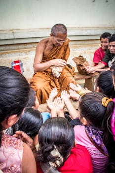An Asian monk in orange robes distributes food to eager children outdoors.