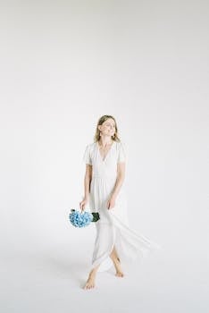 Elegant woman in white dress holding a blue hydrangea bouquet against a minimalist background.