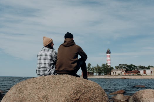 pexels-photo-5081501-5081501 A couple sits on a rock by the sea, gazing at a lighthouse under a clear sky.