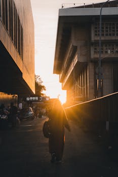 Person walking at sunset between buildings, capturing urban life and warm light in a bustling cityscape.