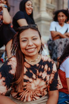 A smiling woman with long hair and eyeglasses sits outdoors, wearing a tie-dye shirt. Urban setting.