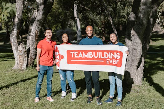 Group of four diverse adults holding a team building banner in a sunny park.